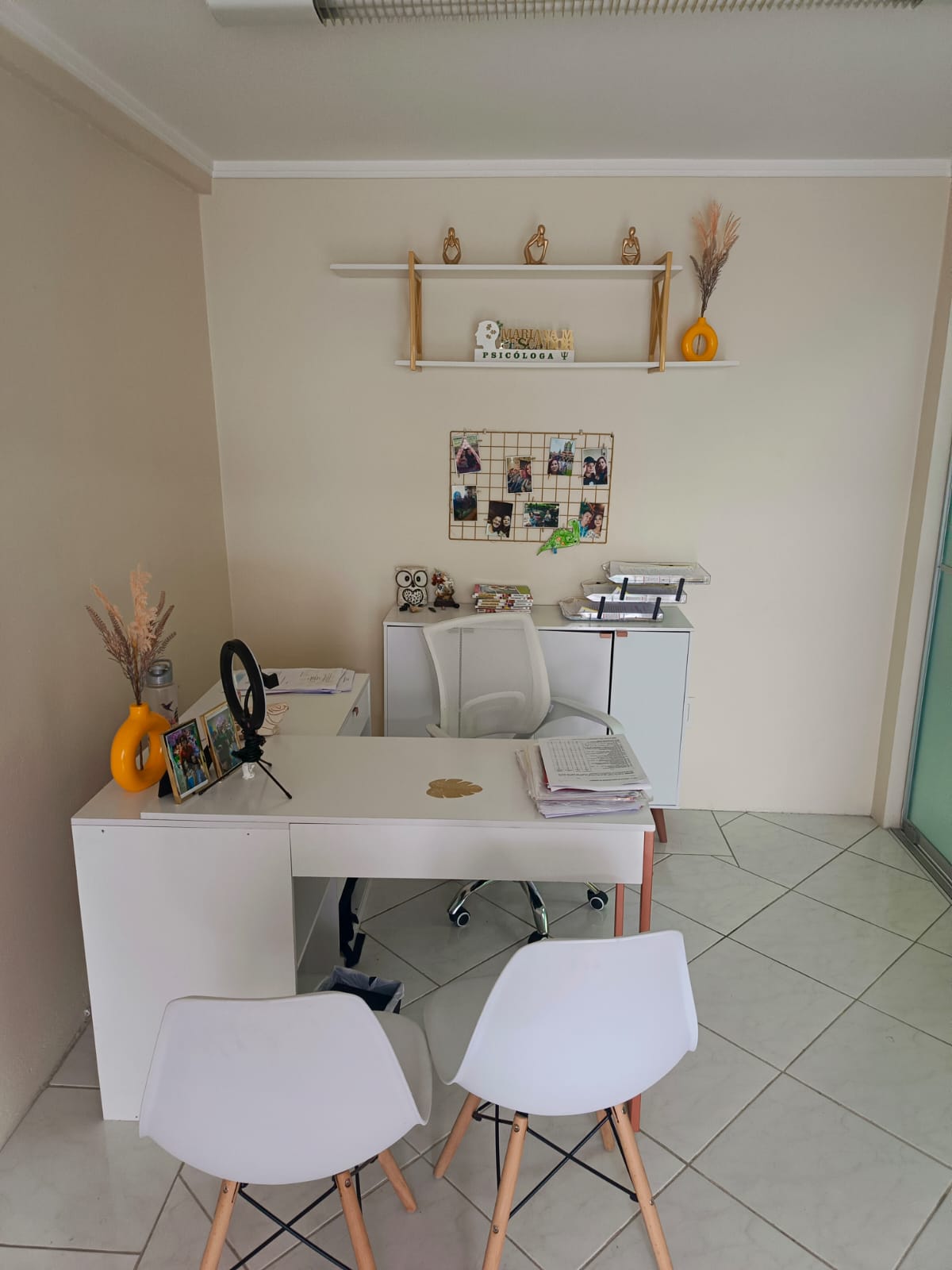 Escritório da psicóloga com mesa branca, cadeiras e prateleira decorada